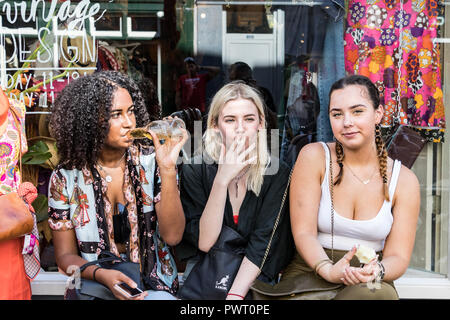 Amsterdam, Netherlands - July 23th, 2018: Three young women, back, blond and brunette, sitting outsiide in the street eating, smoking and drinking bee