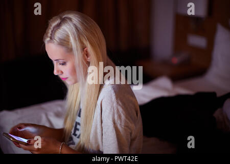 Front view of a sad teen checking phone sitting on the floor in the living room at home with a dark background. Stock Photo