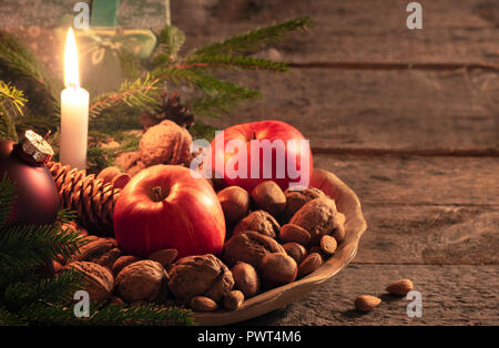 Christmas scenery with a wooden platter full of walnuts, hazelnuts and apples, green fir twigs and a gift box, on a vintage table, at candlelight. Stock Photo
