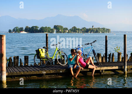 Cyclists resting on jetty, Gstadt am Chiemsee, behind Frauenchiemsee Island, Chiemgau, Upper Bavaria, Bavaria, Germany Stock Photo