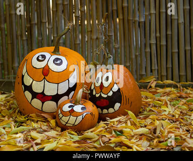 Three smiling painted jack-o-lantern pumpkins, a large, medium and small, sitting among dried leaves with bamboo in the background. Stock Photo
