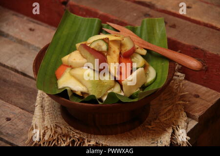 Rujak Buah, the Indonesian fruit salad with spicy coconut sugar dressing. Plated on a shallow wooden bowl lined with banana leaf, Stock Photo