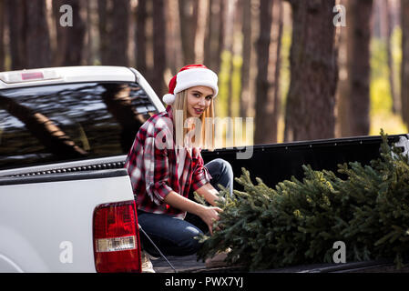 happy girl in santa hat sitting on pickup truck with fir tree for christmas Stock Photo