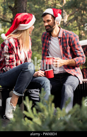 happy couple in santa hats holding red cups while sitting on pickup truck with fir tree for christmas in forest Stock Photo