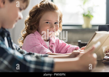 adorable little schoolgirl looking at camera while reading book with brother using tablet blurred on foreground Stock Photo