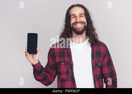 Portrait of positive handsome young adult businessman in red checkered shirt and long curly hair standing and showing smartphone with toothy smile. In Stock Photo