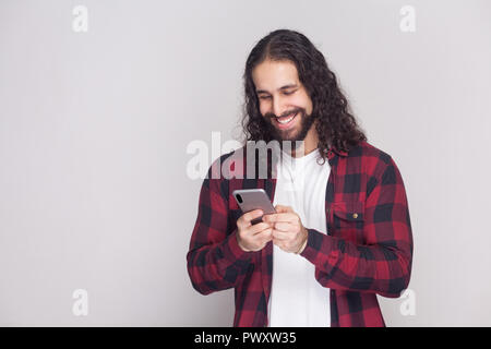 Cheerful handsome young man in red checkered shirt and long curly hair standing, using smartphone and texting message with toothy smile. Indoor, studi Stock Photo