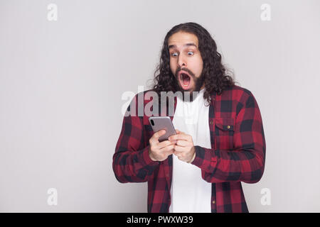 Shocked handsome young man in red checkered shirt and long curly hair and beard standing, using smartphone and watching video with amazed face. Indoor Stock Photo