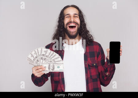 Young happy businessman in red checkered shirt and long black curly hair standing, showing fan of dollars, phone screen with amazed surprised face. In Stock Photo