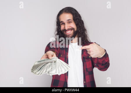 Portrait of satisfied happy young businessman in red checkered shirt and black long curly hair standing, giving and pointing with finger fan of money. Stock Photo