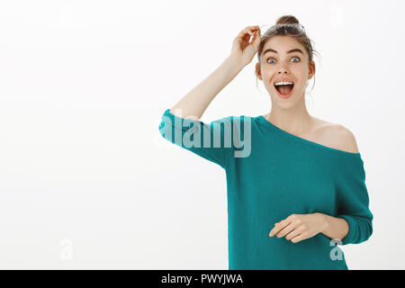 Wow, did not expect see you here. Surprised excited good-looking girl with bun hairstyle, smiling joyfully and taking off glasses while being stunned and amazed, standing happy over gray wall Stock Photo
