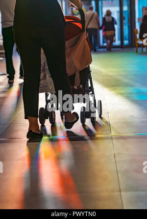 Young mother walking with baby carriage in the shopping center, from the back, evening sun shine reach inside through the swing door. Stock Photo