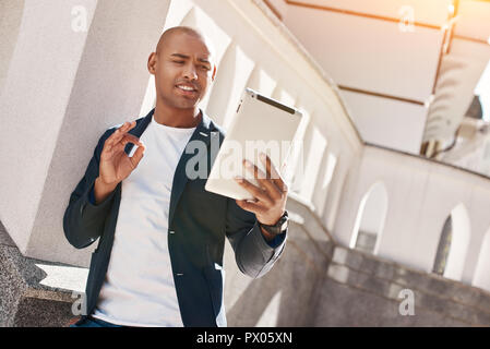 Online communications. Young man standing on the city street having video chat on digital tablet showing ok sign smiling joyful Stock Photo