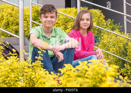 Happy teenage boy and girl smiling while sitting on the stairs outdoors. Young sister and brother teens looking at camera. Stock Photo