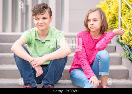 Happy teenage boy and girl smiling while sitting on the stairs outdoors. Young sister and brother teens. Stock Photo
