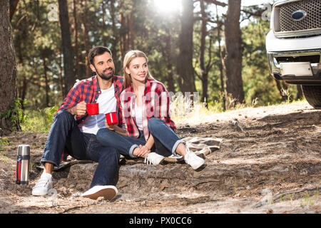 happy young couple sitting on ground with coffee cups near car outdoors Stock Photo