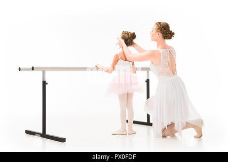 rear view of adult female teacher in tutu helping little ballerina exercising at ballet barre stand isolated on white background Stock Photo