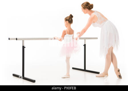 rear view of adult female teacher in tutu helping little ballerina exercising at ballet barre stand isolated on white background Stock Photo