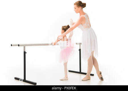 side view of female teacher in tutu helping little ballerina practicing at ballet barre stand isolated on white background Stock Photo