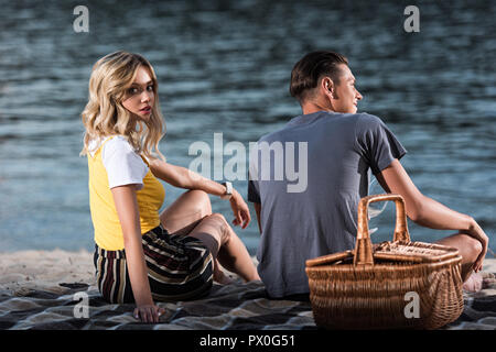 young couple spending time at picnic on river beach in evening, girlfriend looking at camera Stock Photo