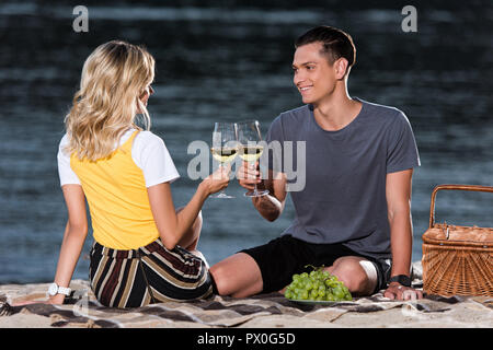side view of young couple clinking with wineglasses at picnic on river beach in evening Stock Photo