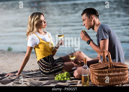 young couple drinking wine and eating grapes at picnic on river beach in evening Stock Photo