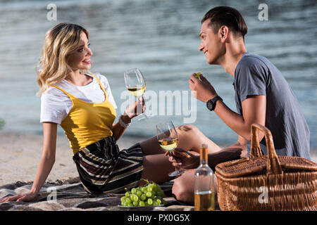 smiling young couple drinking wine and eating grapes at picnic on river beach in evening Stock Photo