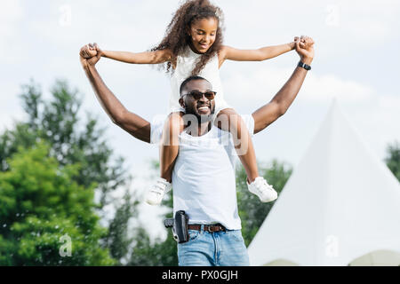 happy african american police officer with gun holding daughter on shoulders in amusement park Stock Photo