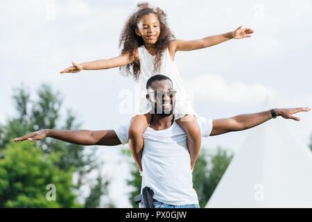 african american police officer with gun holding daughter on shoulders and they pretending flying in park Stock Photo