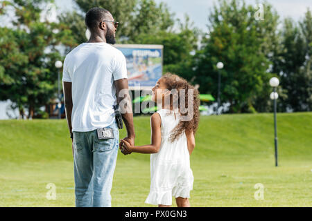 rear view of african american police officer and daughter holding hands and walking in amusement park Stock Photo