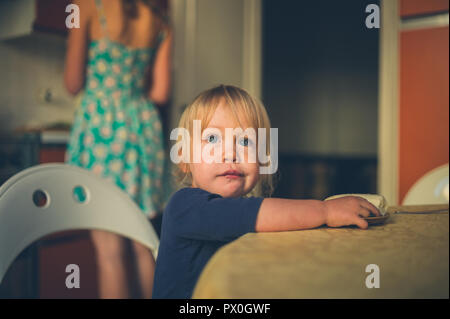 A little toddler is sitting in the kitchen with his mother cooking in the background Stock Photo