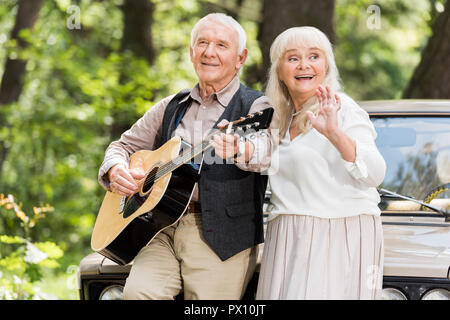 beautiful senior woman smiling with man playing guitar leaning on car Stock Photo