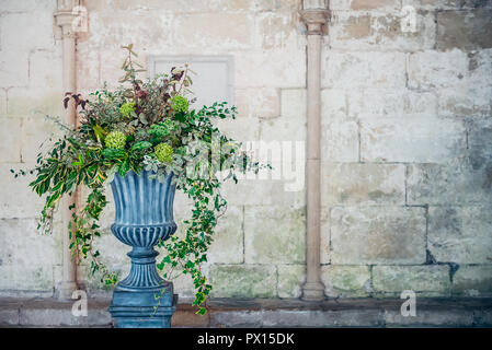 Stone vase planter with fustian green flowers and cascading green ivy and leaves. Grey cobble stones. Gorgeous design in front of medevial wall with c Stock Photo