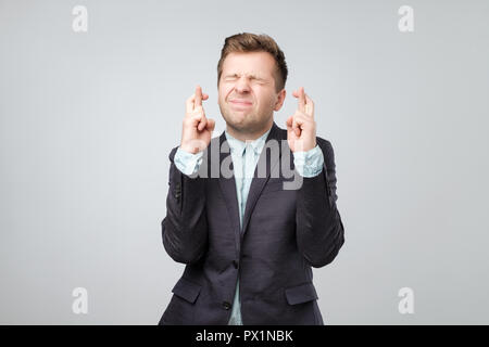 Handsome young man in suit looking at camera and keeping fingers crossed wishing some luck while standing against grey background Stock Photo