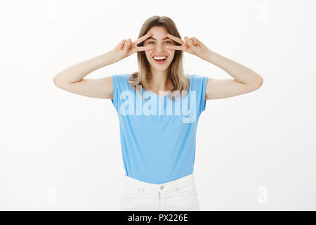 Portrait of happy emotive good-looking woman in trendy t-shirt, showing disco gesture or victory over eyes and smiling broadly, having fun, spending time with friends, being in playful and joyful mood Stock Photo