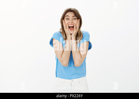 Indoor shot of happy emotive european female fan in blue t-shirt, screaming and smiling, holding palms on cheeks, gazing at camera while seeing favorite singer, having fun on music concert Stock Photo