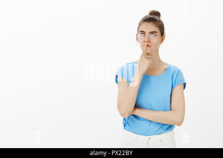 Need to find solution quickly. Portrait of determined serious-looking girl in bun and glasses, looking aside with focused expression, holding finger on lip while thinking or making up plan Stock Photo