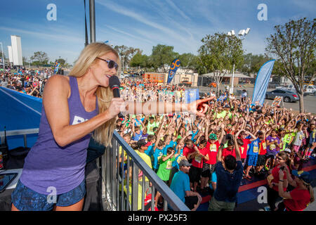 From an overhead platform, a blonde hostess with a microphone tells 10-year-old foot multiracial runners to stretch out before the race begins at a community track event in Costa Mesa, CA. Stock Photo