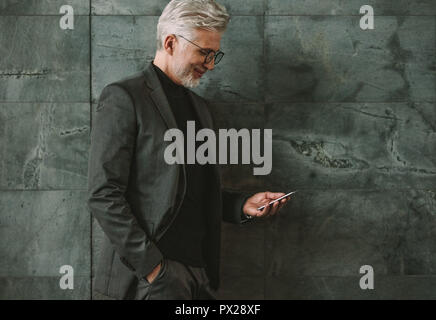 Mature businessman leaning to a wall and looking at cellphone. Senior businessman using mobile phone on grey background. Stock Photo