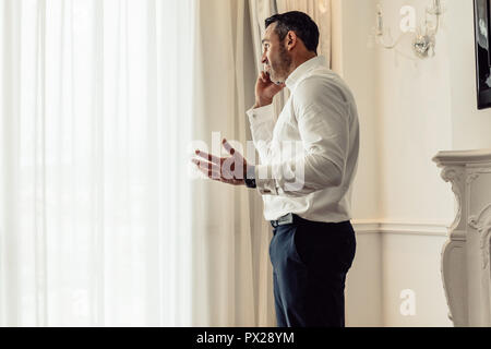 Side view of businessman talking on mobile phone while standing in hotel room. CEO having an important discussing over phone. Stock Photo