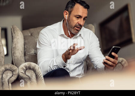 Mature businessman wearing earphones talking on mobile phone. Man on business trip sitting in hotel room and making phone call. Stock Photo