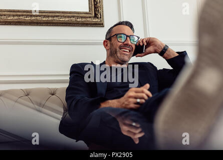 Portrait of relaxing mature businessman sitting on couch and talking on phone. Man in suit relaxing in hotel room and making phone call. Stock Photo