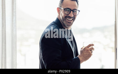 Stylish mid adult man looking away and smiling with a drink in hand. Businessman having a drink in hotel room. Stock Photo