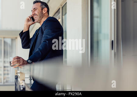 Happy mature businessman standing in hotel room balcony and making a phone call. Smiling man in suit talking on mobile phone from hotel room. Stock Photo