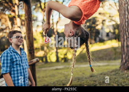 Girl hanging upside down in a park holding a heart shaped candy lollipop. Smiling boy standing in a park while looking at his girlfriend who is hangin Stock Photo