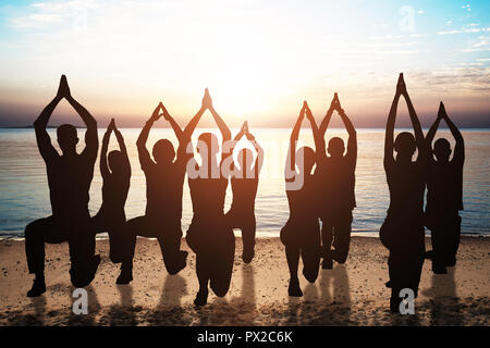 Silhouette Of People Doing Yoga On Sandy Beach At Sunset Stock Photo