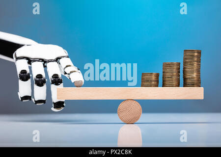 Robot Balancing Stacked Coins With Finger On Wooden Seesaw Against Blue Background Stock Photo