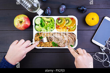 Top view showing hands eating healthy lunch with bulgur, meat and fresh vegetables and fruit on a wooden table. Fitness and healthy lifestyle concept. Stock Photo