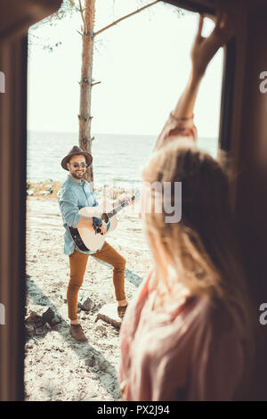 selective focus of girl standing in door of campervan while man playing guitar Stock Photo