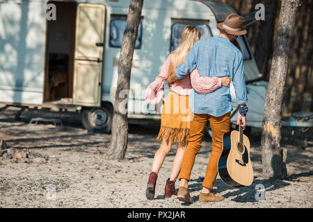 rear view of hippie couple hugging and walking with guitar near trailer Stock Photo